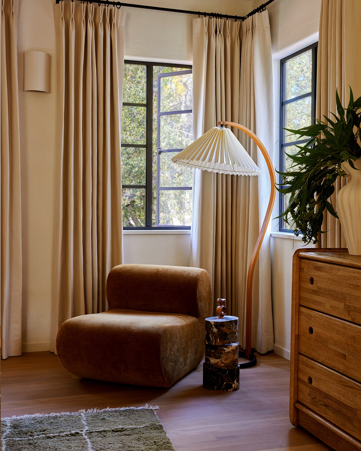 A styled corner with floor-length beige curtains, a brown upholstered lounge chair, a curved floor lamp, a wooden dresser, and a potted plant near tall windows.