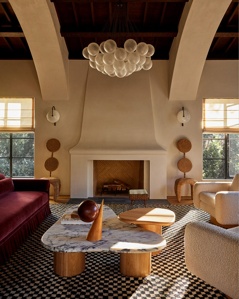 A living room interior featuring a distinctive bubble chandelier, a marble-top coffee table, a black-and-white checkered rug, a deep red velvet sofa, and a white fireplace with a tiled surround.