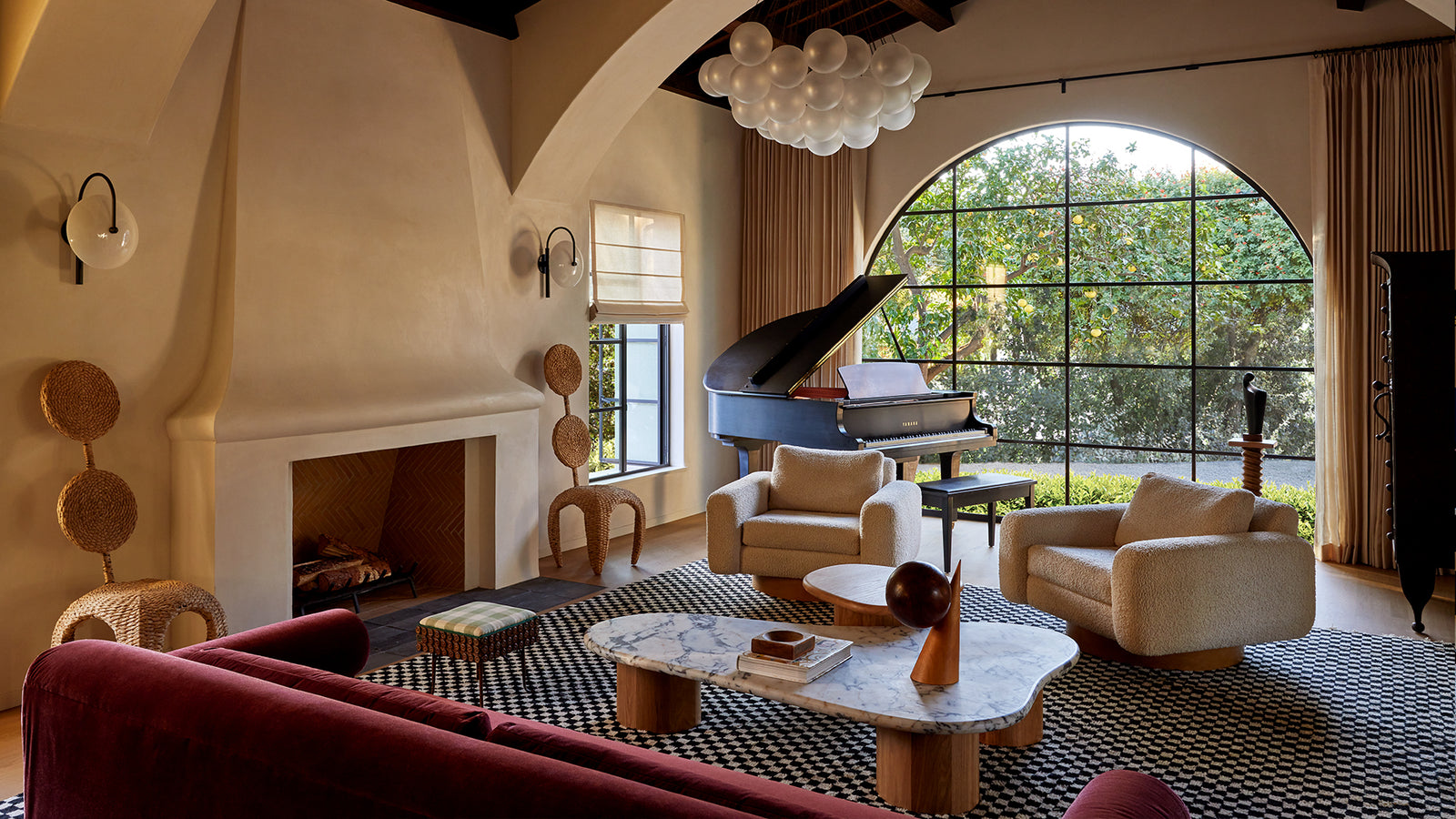 A living room interior featuring a grand black piano, cream armchairs, a red sofa in the foreground, a patterned rug, and a large arched window looking out onto greenery.